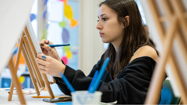 A person painting on a canvas with paintbrushes on the table.