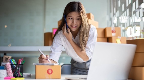 A women working on a computer while on the phone and taking notes on a notepad