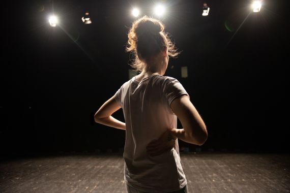 A young girl standing on a stage with their back facing the camera lights on her face