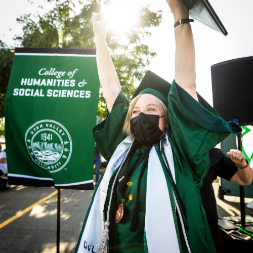 female student dressed in green cap and gown, arms raised in triumph