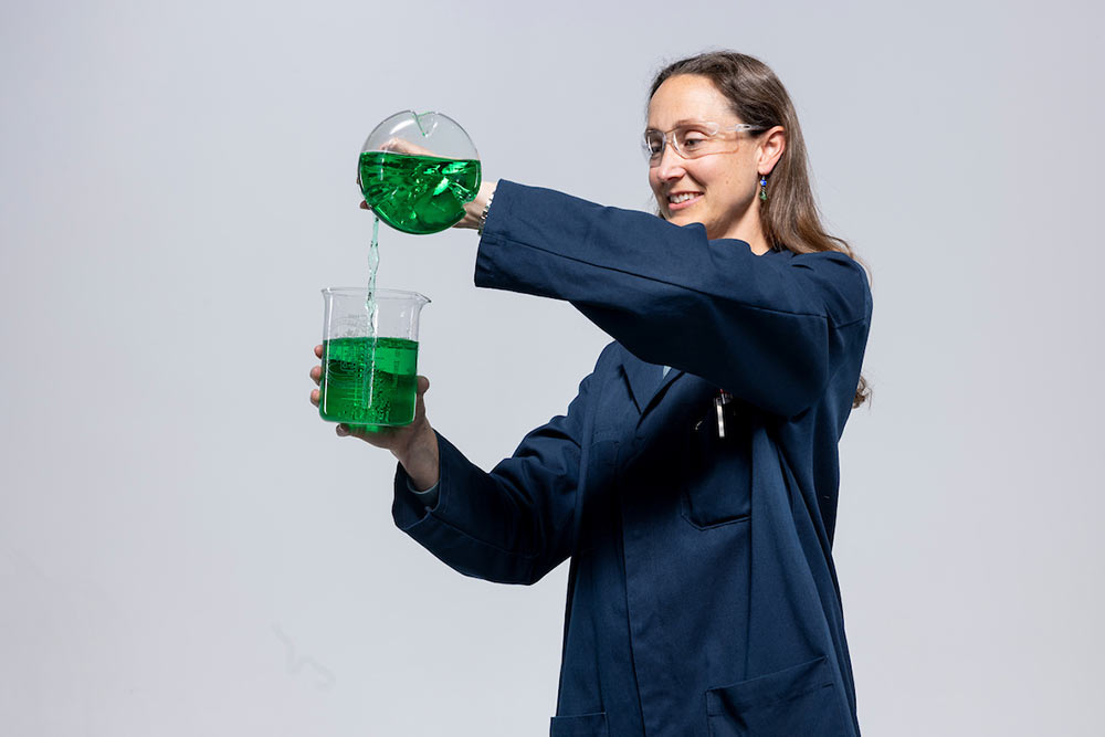 Female wearing a lab coat, pouring liquid from one beker into another beker.