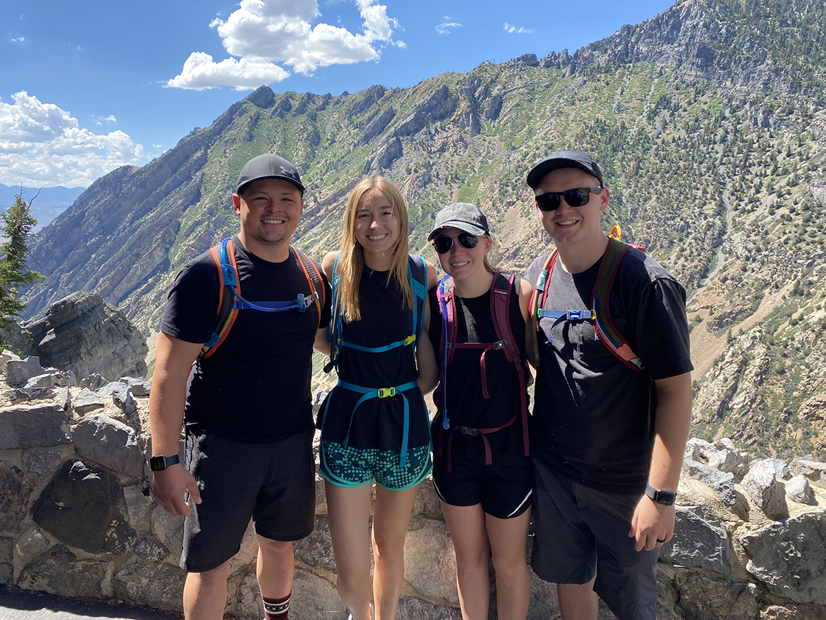 Layton with 2 males and a female in hiking clothes on a mountain interlocking arms and smiling at the camera.