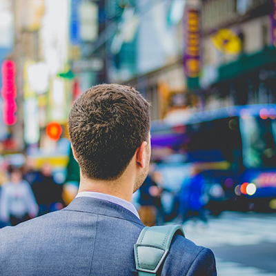 Man with back to camera, in suit with bag on shoulder looking across busy city street