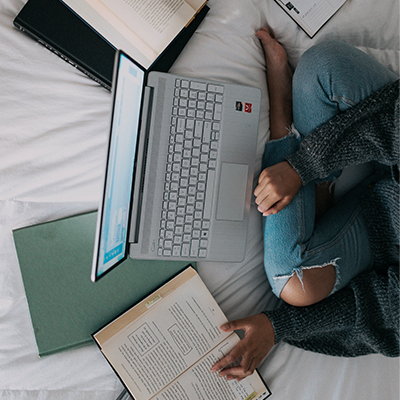 student studying with a computer open on a bed surrounded by books