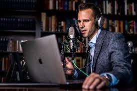 Greg Jackson sitting behind MacBook while wearing headphones and talking into podcast microphone