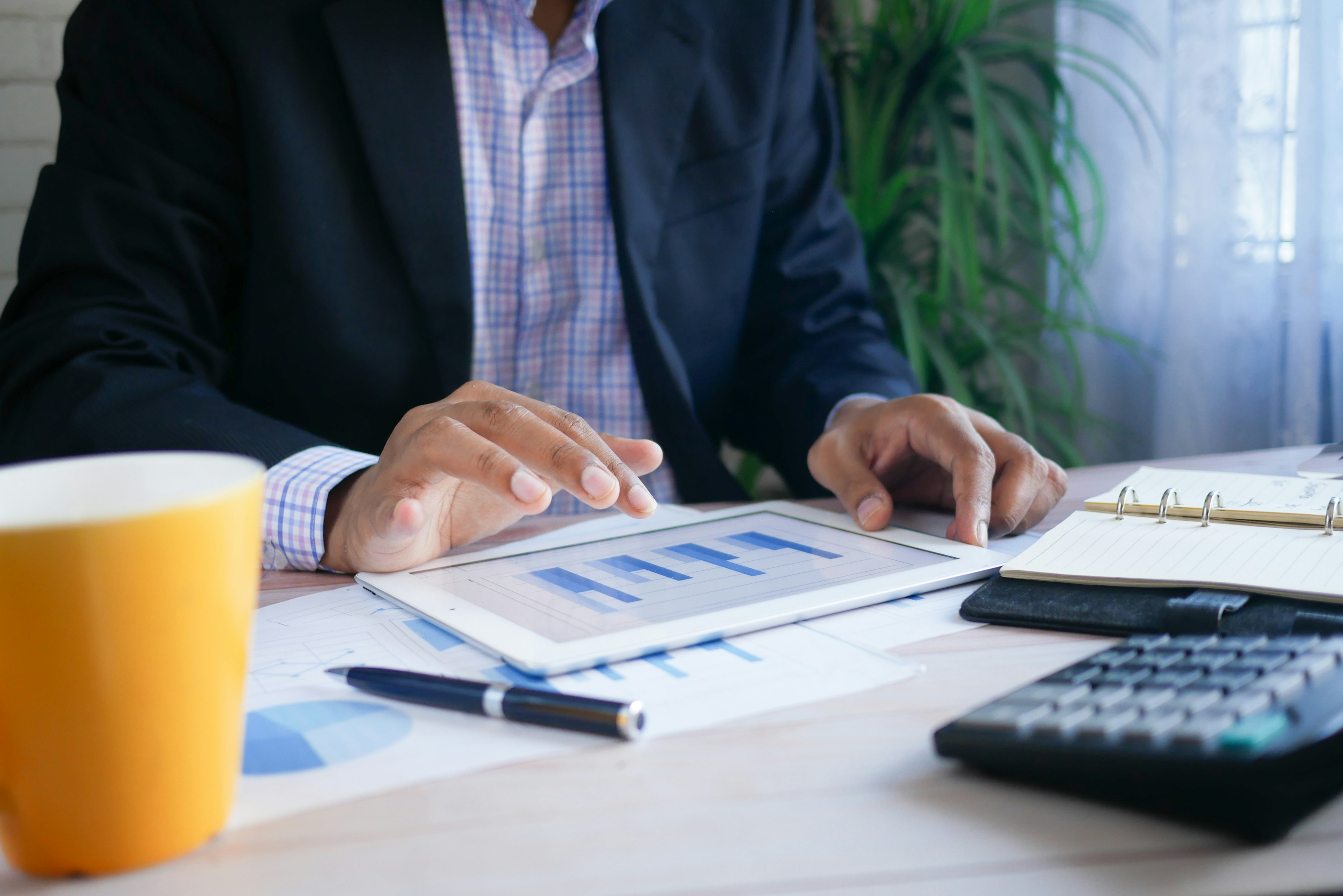 A person in a blazer uses a tablet showing a bar graph on a wooden table with a yellow cup, a pen, printed documents, a notebook, and a calculator.