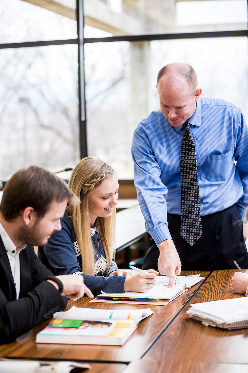 A teacher assists two students at a table in a bright classroom, pointing at an open book while one student writes in a notebook.