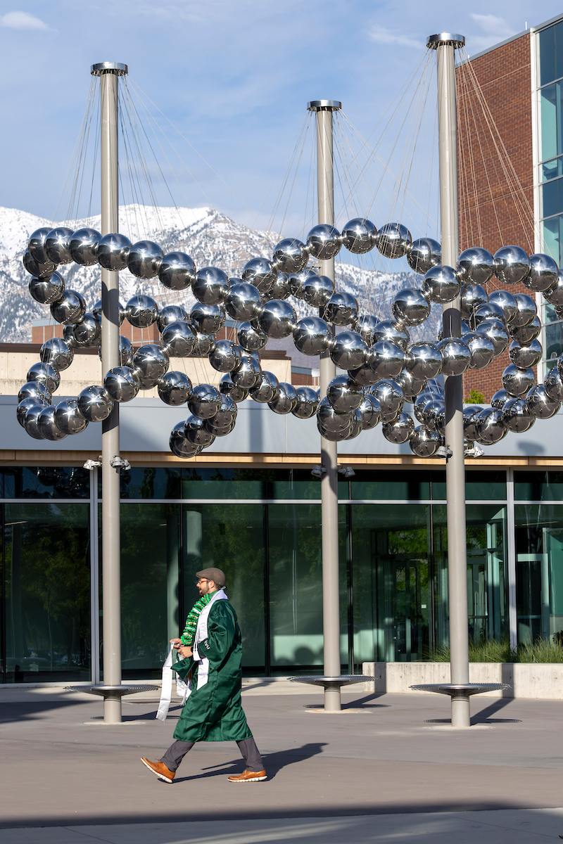 UVU Grad walking past an abstract silver sculpture and UVU building, with mountains in the background.