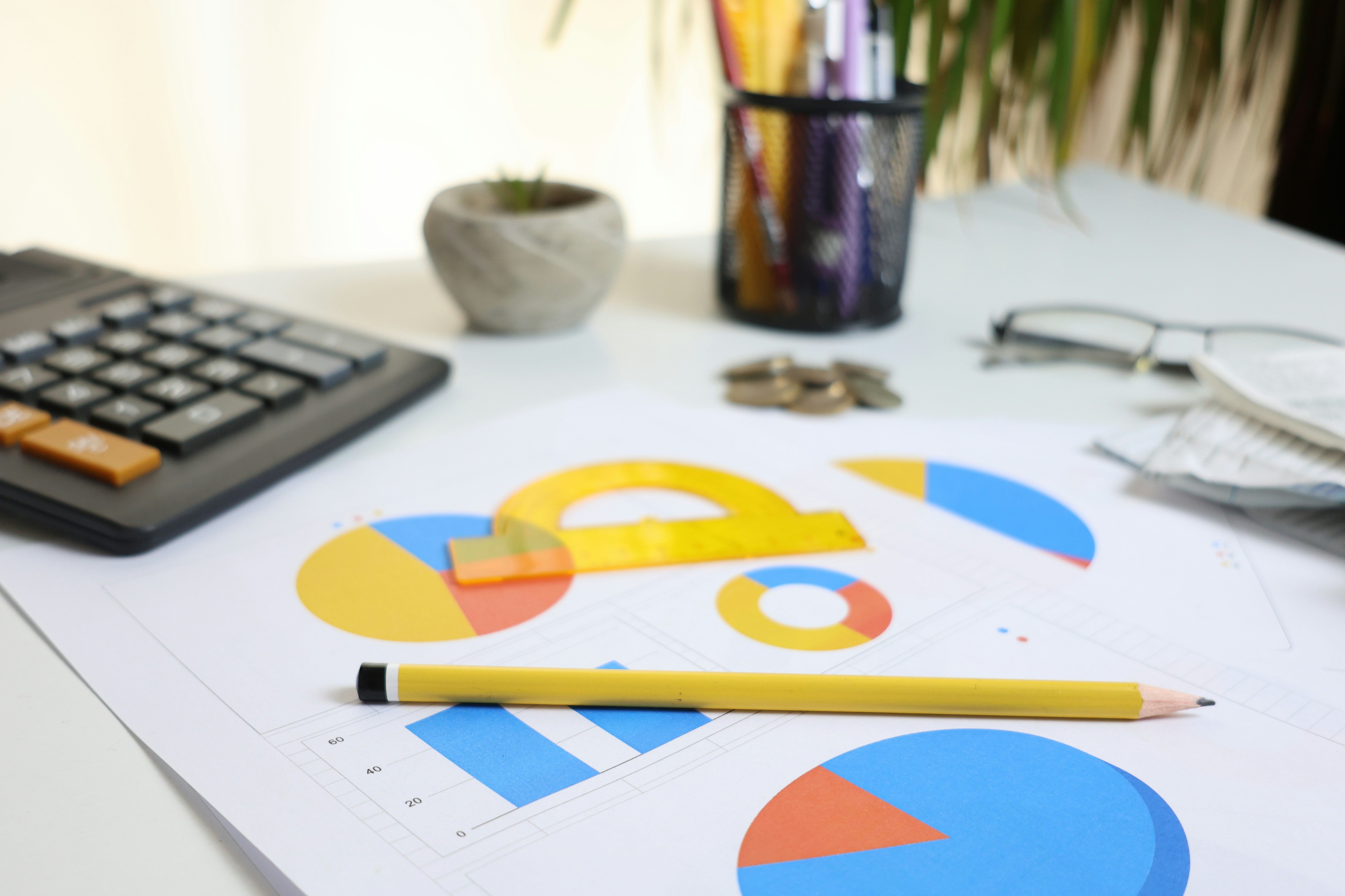 A workspace featuring printed graphs and charts, a yellow pencil and ruler, a calculator, a small plant, and other stationery.