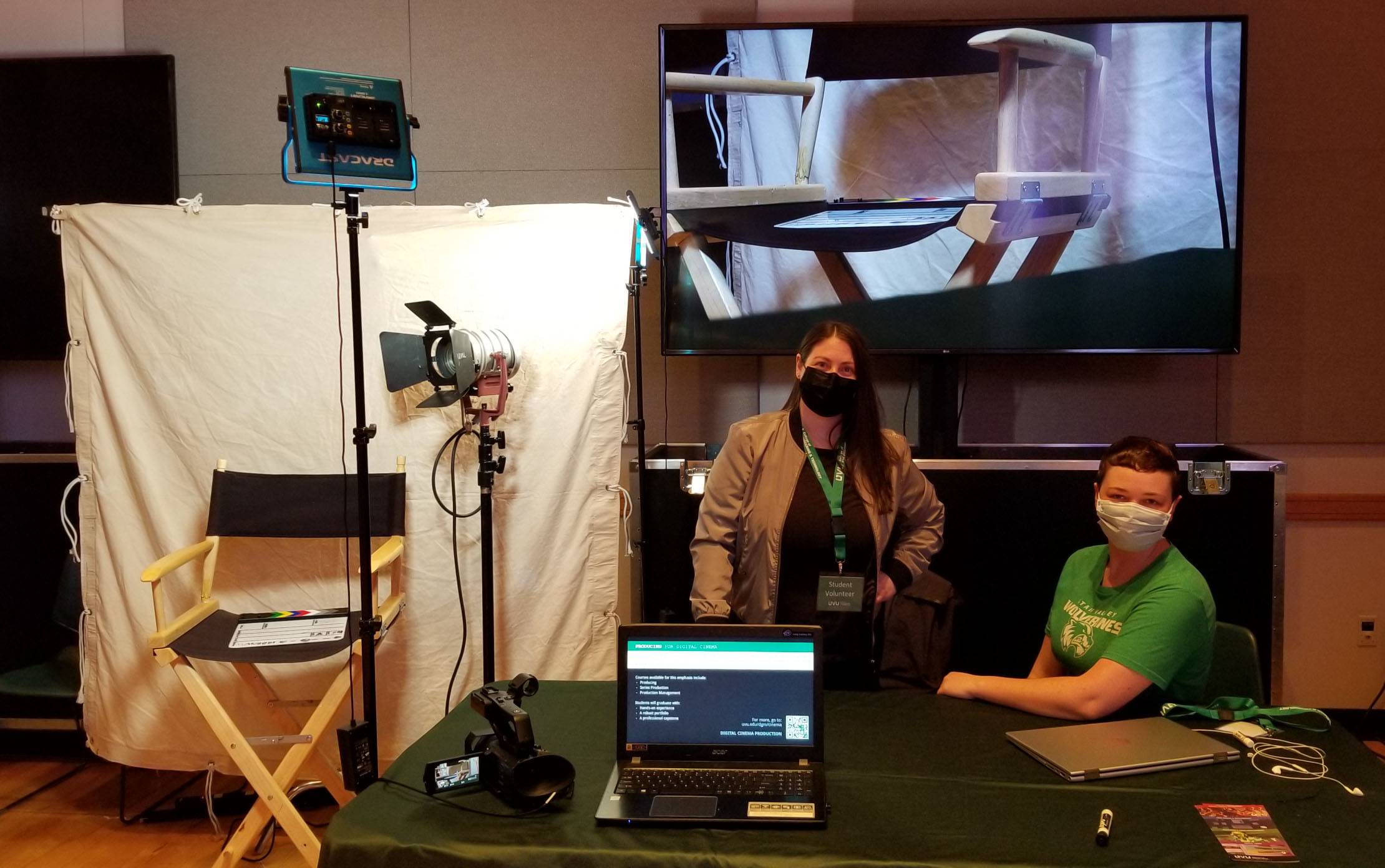 Female cinema students Alysha Aldred standing on left and Ariana Wray sitting on right in front of large screen showing the director's chair to their left surrounded by lighting in front of a drop cloth a video camera and laptop computers are on the table.