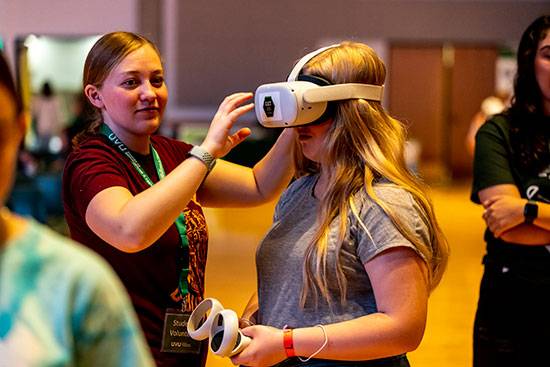 Female student Mariah Foerster blonde wearing maroon shirt assisting a girl blonde with a grey shirt with a VR headset on and holding controllers