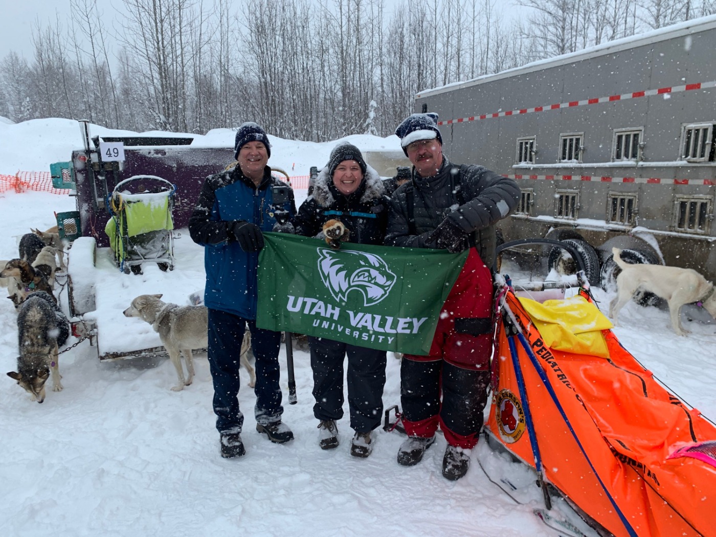 Mike Wisland, Emily Hedrick, and and musher Alan Eischens holding the UVU Flag with sleds and dogs about them.