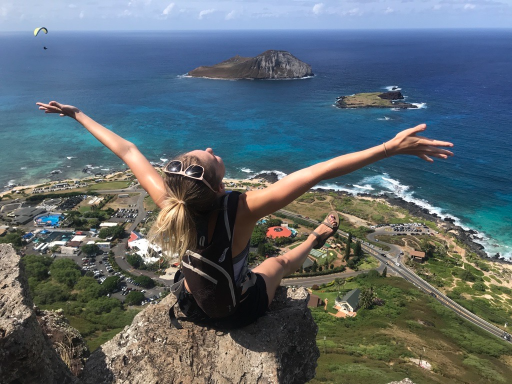 Megan with back to the camera sitting on an outcrop overlooking headland and the water off of Hawaii