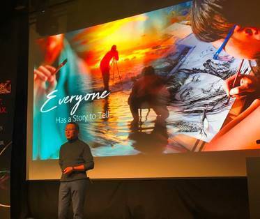 Man on stage with a collage image projected behind him and the words "Everyone has a story to tell"