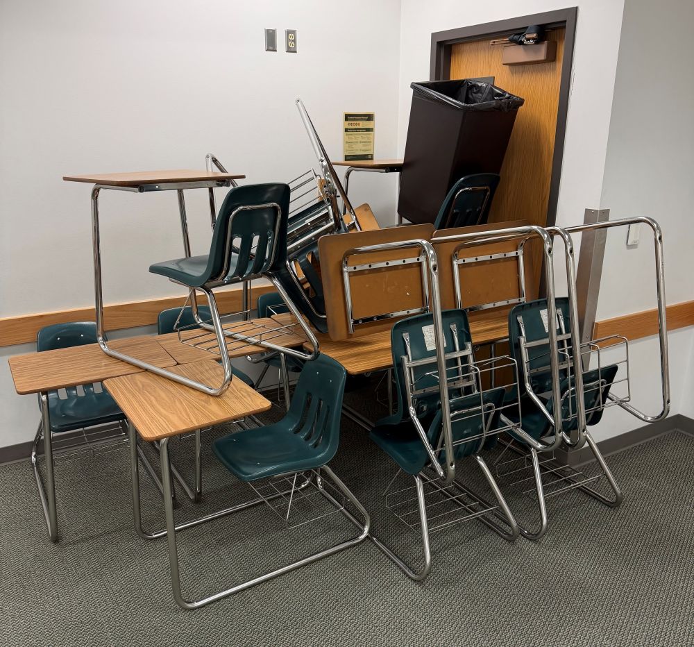 Barricade of chairs in front of a classroom outward opening door. A belt is seen fastened on the door closer.