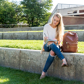 Blond female student sitting outdoors writing with a pen in a notebook