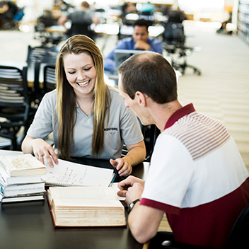 Blonde Female Mentoring Dark haired Male student sitting at a desk with books and papers