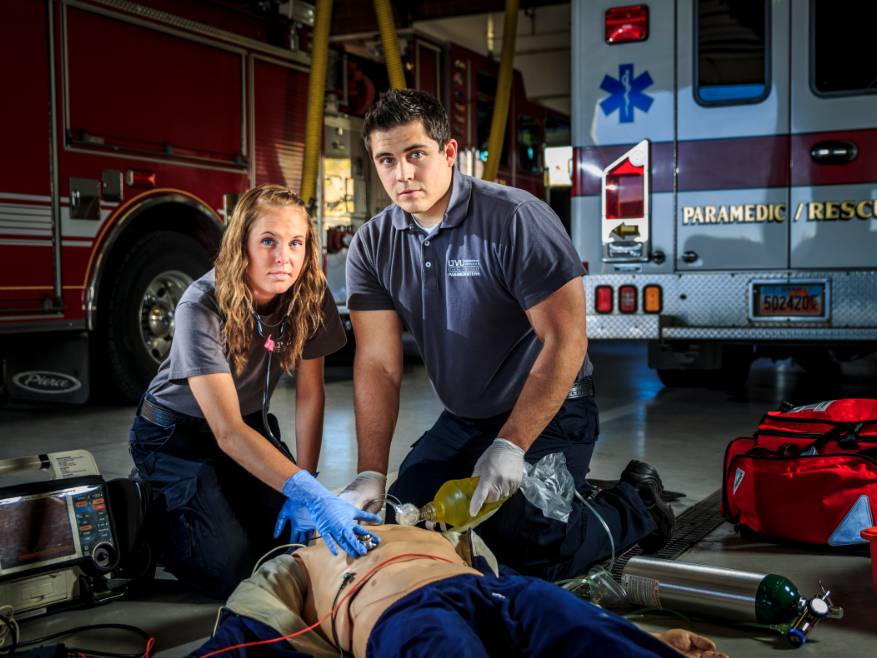 male and female paramedics kneeling on the ground working on a manakin inside an emergency vehicle hanger