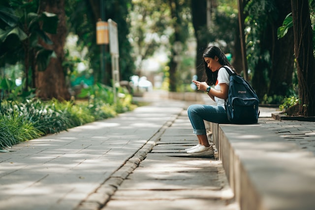 girl with backpack sitting on a curb looking at her phone
