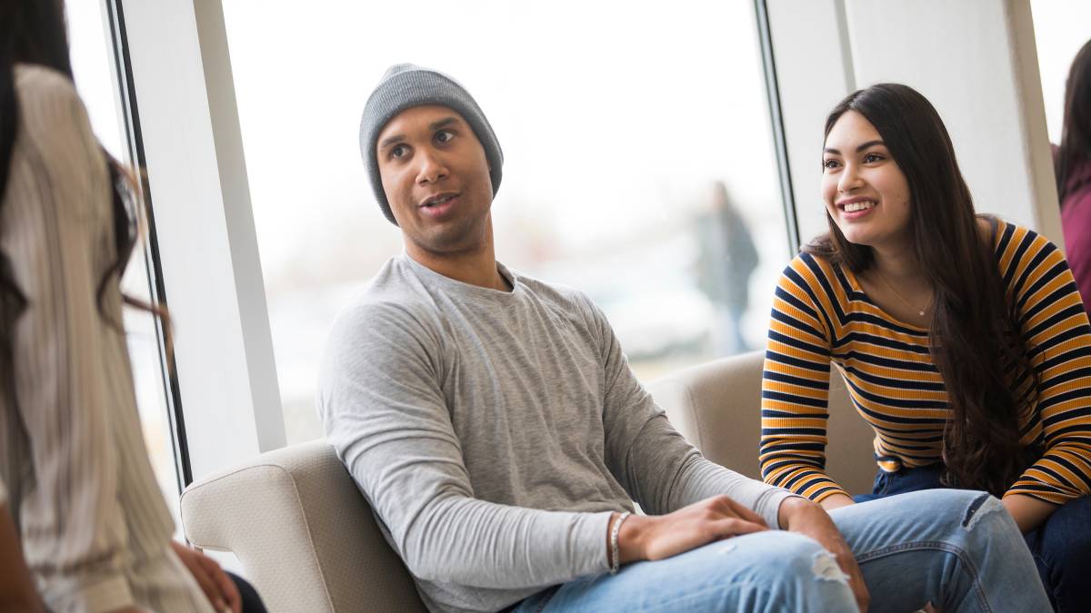 Two students sitting on a couch while talking to another student.