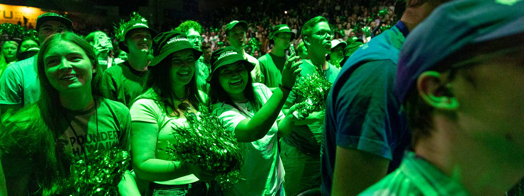 A green light shines on a big group of people with some of them holding green pom-poms.