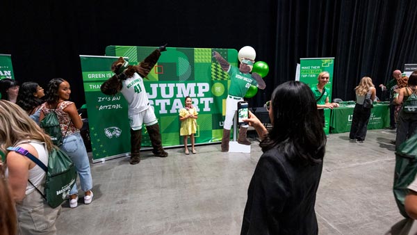 A young girl posing with Willy the Wolverine in front of a sign that says Jump Start while a woman takes a picture of them.