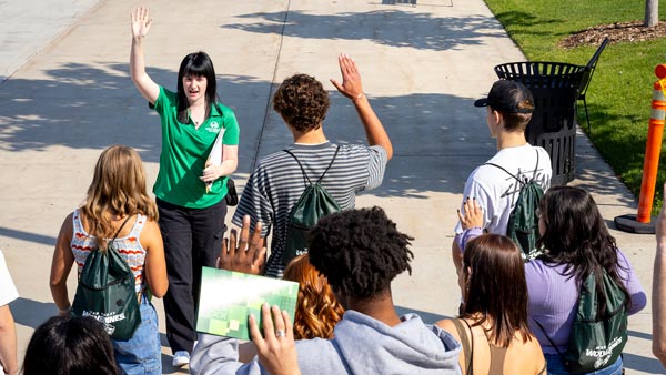 A group of new students follow a woman in a light green shirt, both she and some of the students are raising their hands.