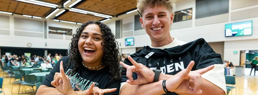 A woman and a man wearing UVU attire and holding up the Wolverine hand sign.