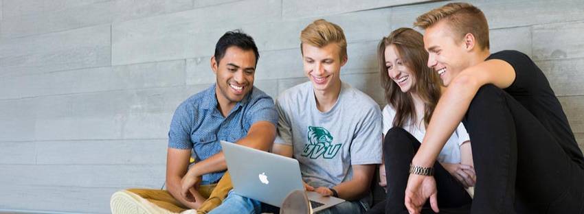 Four UVU students sitting up against a brick wall looking at a laptop together.