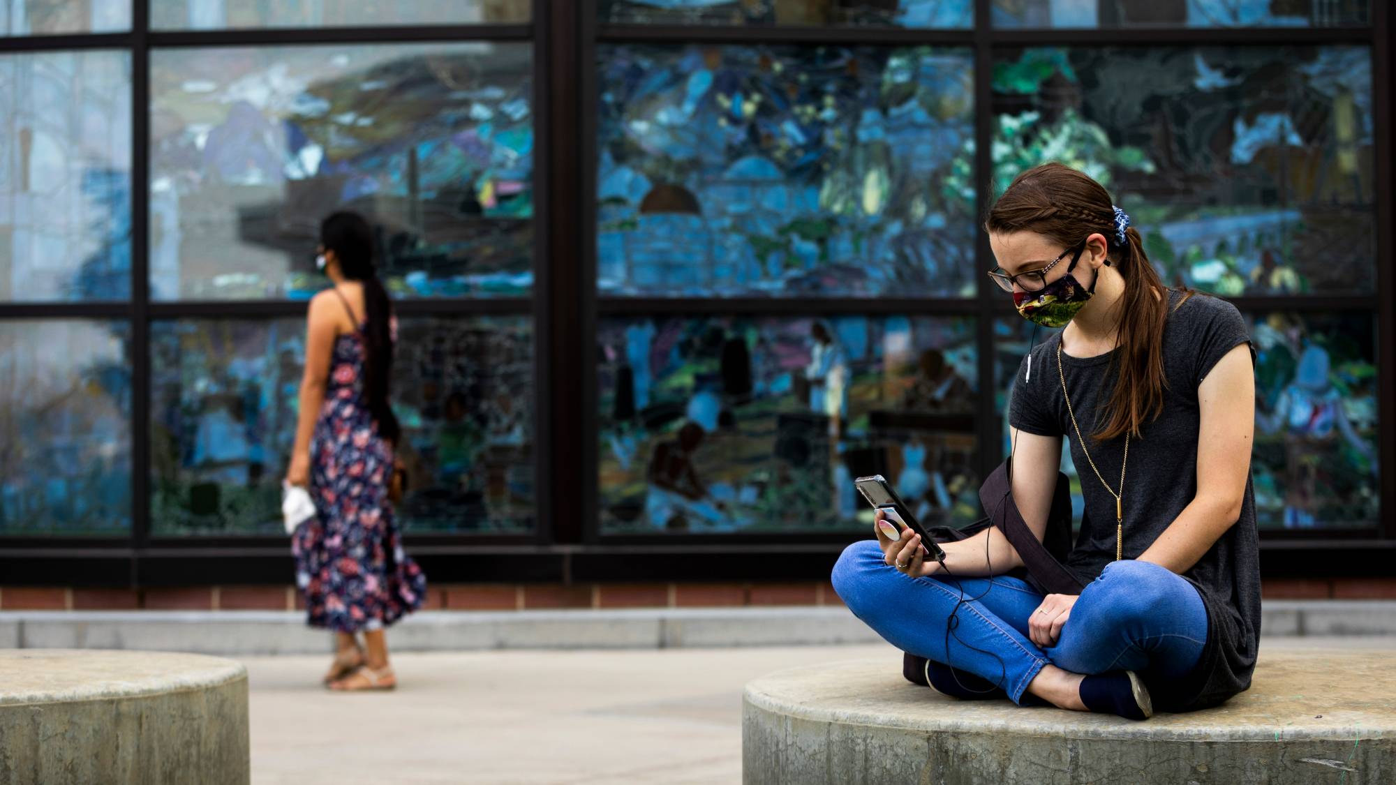 Seated female student wearing a face mask, sitting in front of the Roots of Knowledge windows.