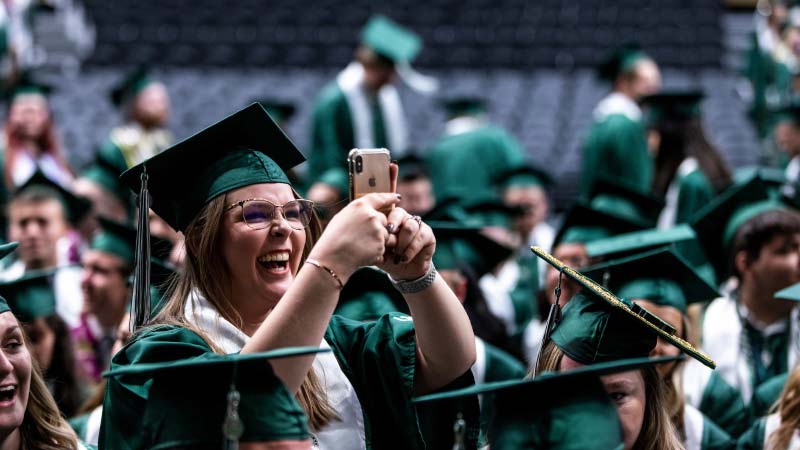 Graduation Checklist A UVU graduate standing, taking a picture on a cell phone, in the middle of other seated UVU graduates.