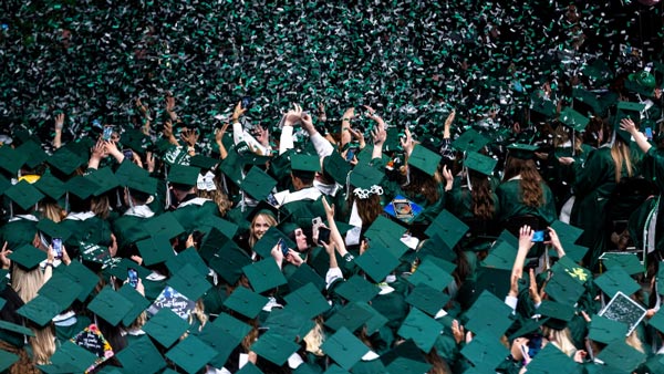Graduates dress in UVU graduation gowns cheer as green and white confetti falls around them