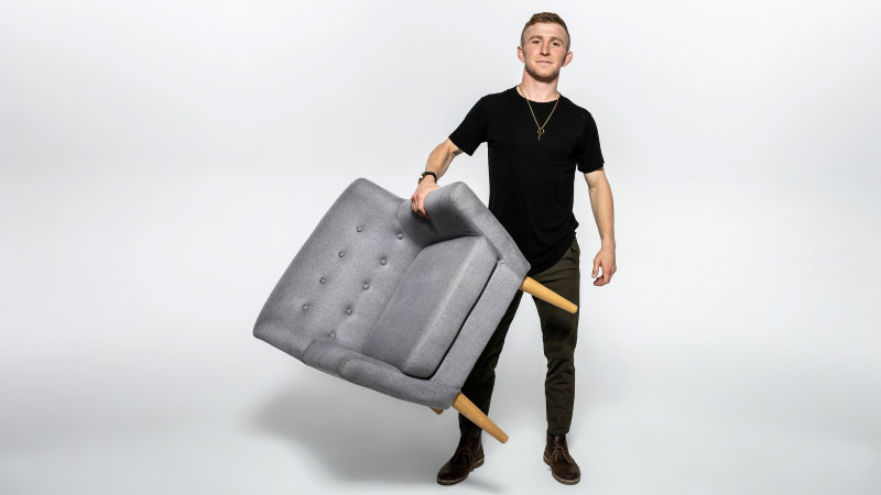 What To Bring A man standing, holding a chair against his body with one hand.