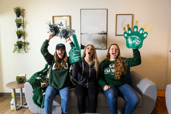 Three women dressed in UVU attire, sitting on a couch, cheering