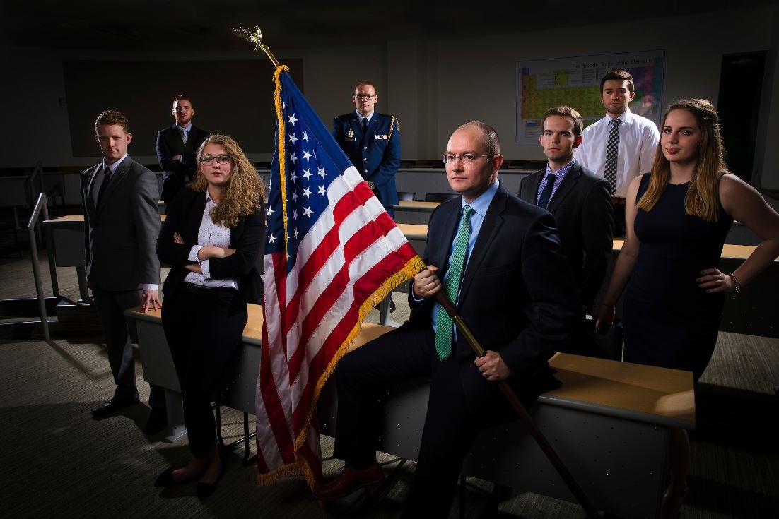 Picture of students and professor sitting in a room gathered around an American Flag