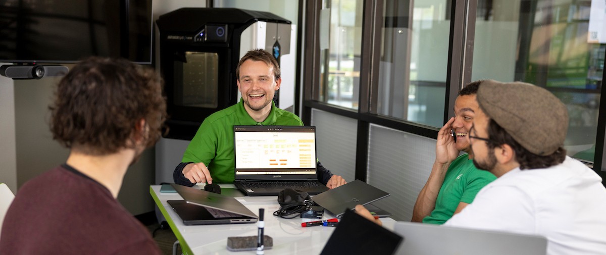 students seated around a table, one student is showing code to the group from his laptop