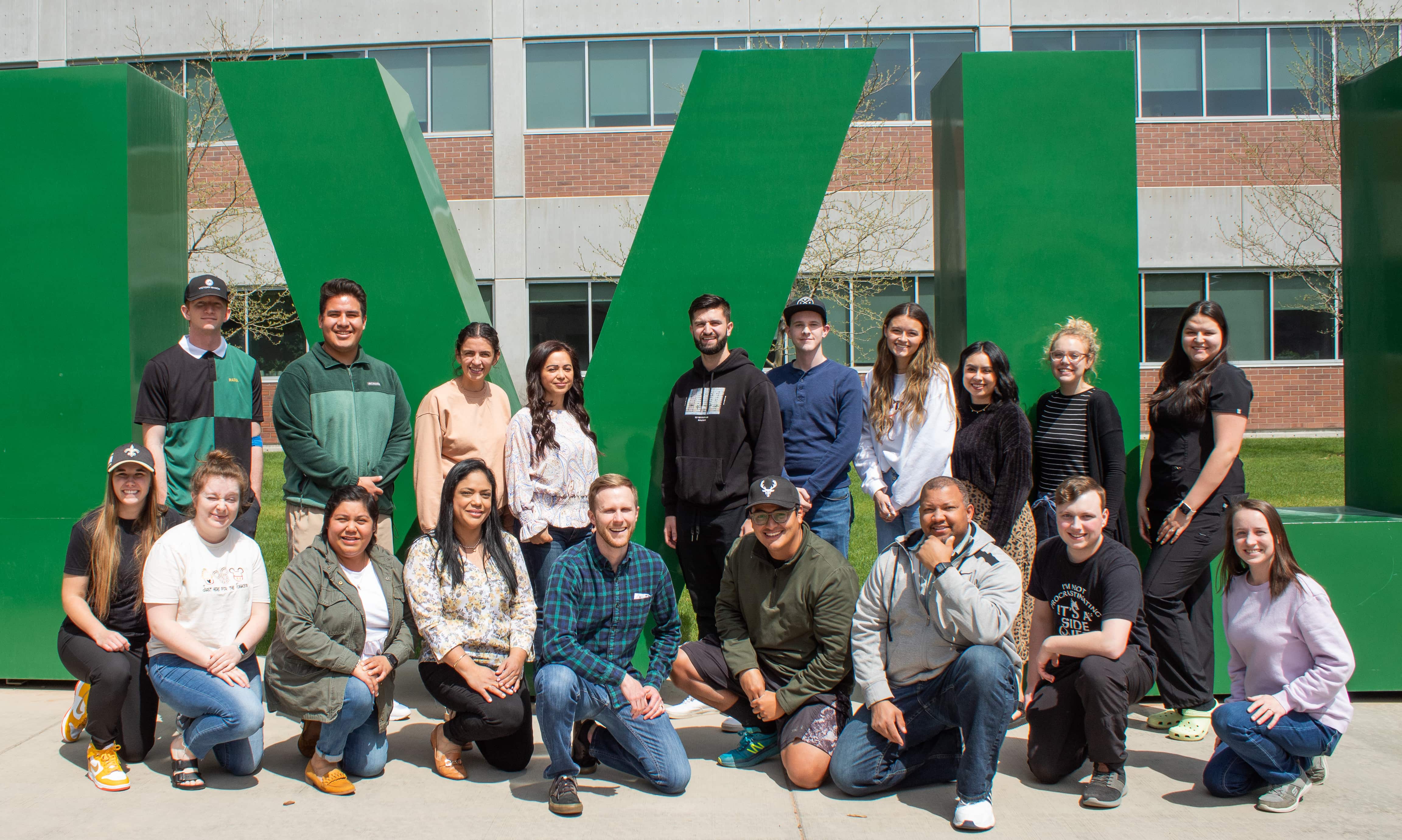 Two rows of students and one professor posing for a picture on a sunny day in front of the giant green UVU letters on the Orem campus