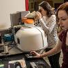 Two women utilize a machine, one views through the machine while the other views information on a computer monitor