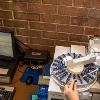 Hands are inserting chemicals into a vial above a machine containing many vials; there is a computer next to it with data displaying on the screen