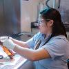 A woman sits at a desk wearing safety goggles using a tool on a glass bottle