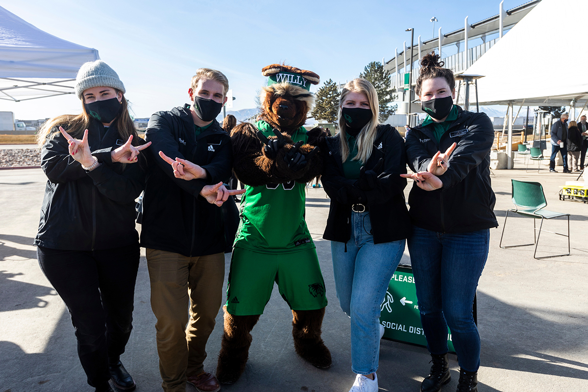 Four event ambassadors stand on either side of Willy the Wolverine mascot and hold up peace signs.