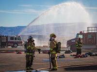 Firefighters standing in front of two firetrucks with one firetruck spraying water.