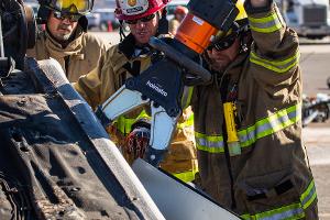 Firefighters gathered around a burnt car using a Hydraulic rescue tool