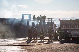 Group of firefighters standing on a building with other firefighters standing on the ground next to a firetruck