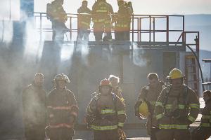 Group of firefighters standing on a building with other firefighters standing on the ground next to a firetruck