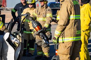 Firefighters gathered around a burnt car using a Hydraulic rescue tool