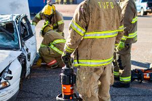 Firefighters gathered around a burnt car using a Hydraulic rescue tool