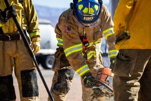 A firefighter cutting plywood with a saw with two others prying it open with tools