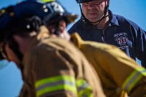 A firefighting instructor supervising a firefighter cutting plywood with a saw