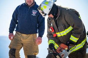 An firefighting instructor supervising while a firefighter cuts plywood with a saw